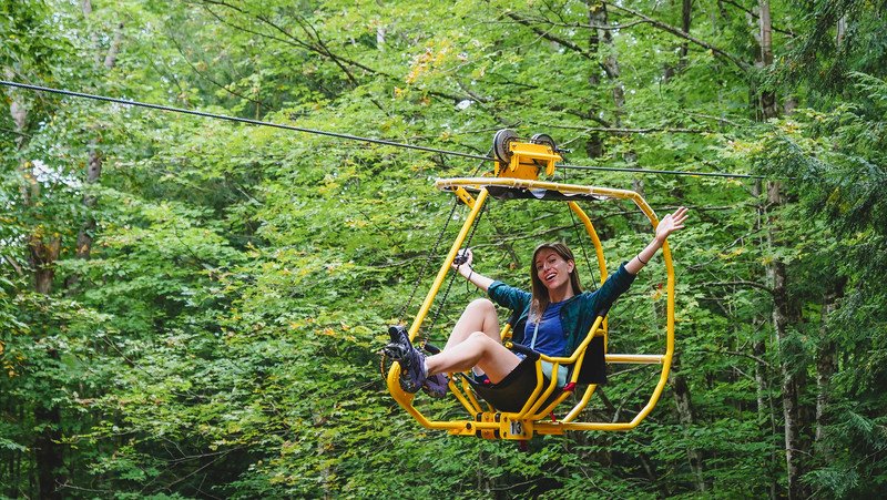 That Backpacker Audrey Bergner joyfully riding the bright yellow Vélo Volant pedal-powered canopy bicycle through the treetops at Au Diable Vert in Sutton, Quebec, surrounded by lush forest in the Eastern Townships