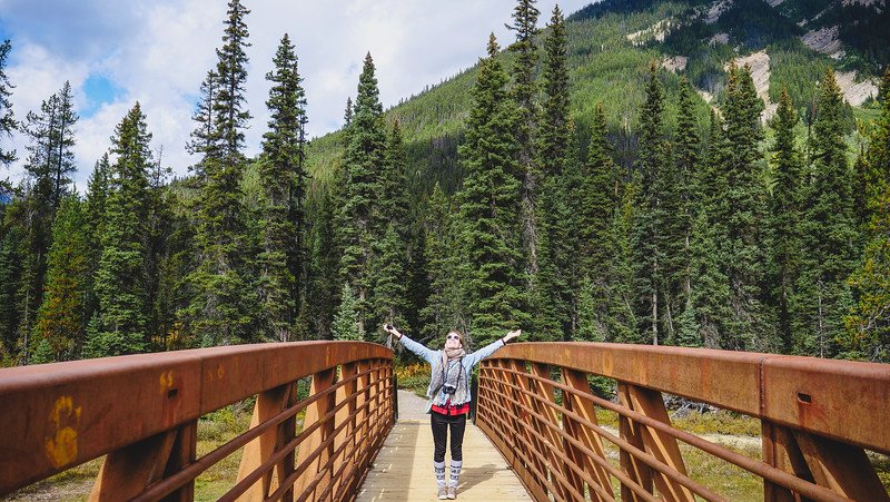 Audrey Bergner from That Backpacker standing joyfully with arms raised on the rust-coloured metal bridge over the Vermilion River, surrounded by dense evergreen forest and mountain slopes on the way to the Paint Pots in the Canadian Rockies