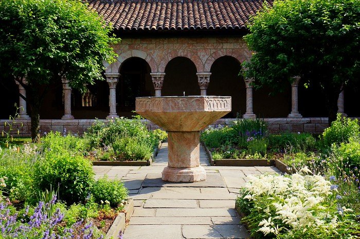 A peaceful medieval-style courtyard garden at The Cloisters in Upper Manhattan, New York City, featuring a central stone fountain surrounded by lush greenery, flowering plants, and arched walkways. This museum offers a tranquil escape from the city