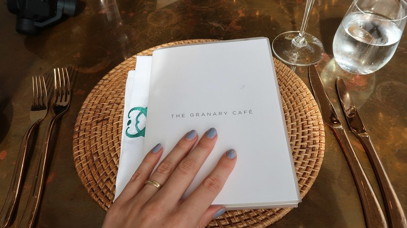 Close-up of a hand holding the elegant menu at The Granary Café in Cape Town, set on a stylish table with woven placemat, gold cutlery, and a glass of water and wine, ready for lunch