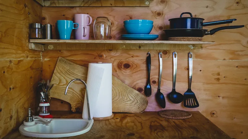 The small kitchen with pump-sink in the tiny house with cups, chopping board and utensils hanging on the wall The small kitchen with pump-sink in the tiny house with cups, chopping board and utensils hanging on the wall