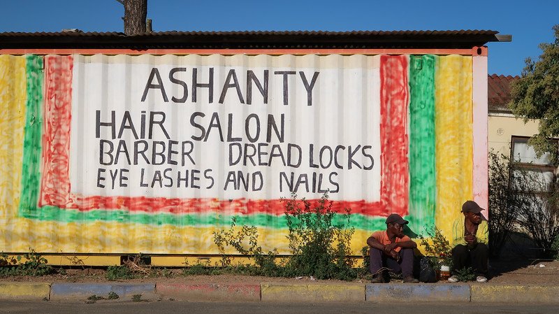 The streets of Kayamandi with the sign Ashanty hair salon, barber, dreadlocks, eye lashes and nails on a red, green and yellow building facade