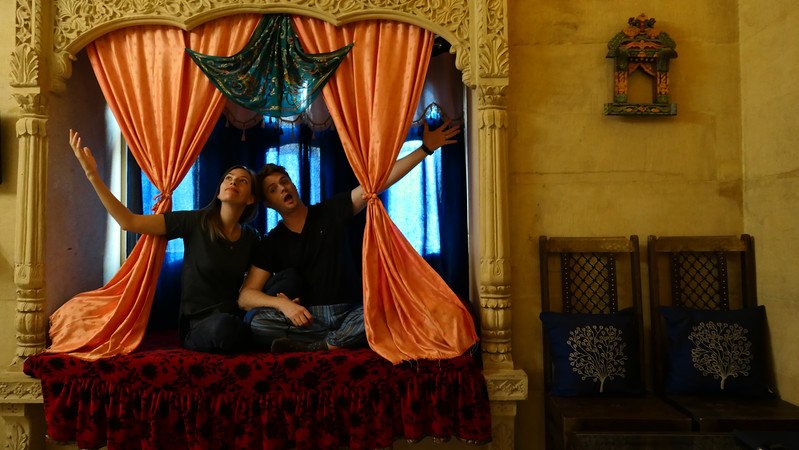 Nomadic Samuel and Audrey Bergner of That Backpacker playfully posing on a carved alcove bed draped with velvet and silk curtains inside a traditional haveli at Tokyo Palace Hotel in Jaisalmer, India