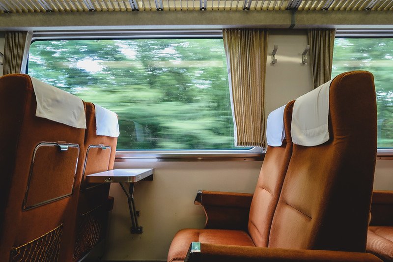 Empty plush train seats with white headrest covers inside a limited express carriage from Tokyo to Nikko, with green countryside scenery blurred outside the window as the train speeds through rural Japan