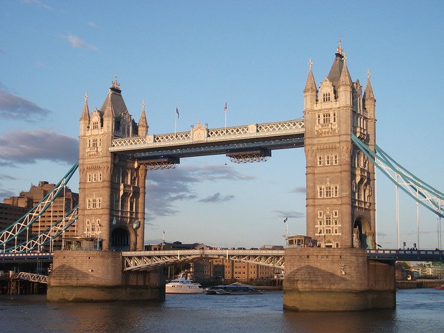 Tower Bridge in London, England, with its twin Gothic-style towers, suspended walkway, and bascule sections over the River Thames, photographed at sunset with boats passing underneath, highlighting one of the city’s most iconic landmarks