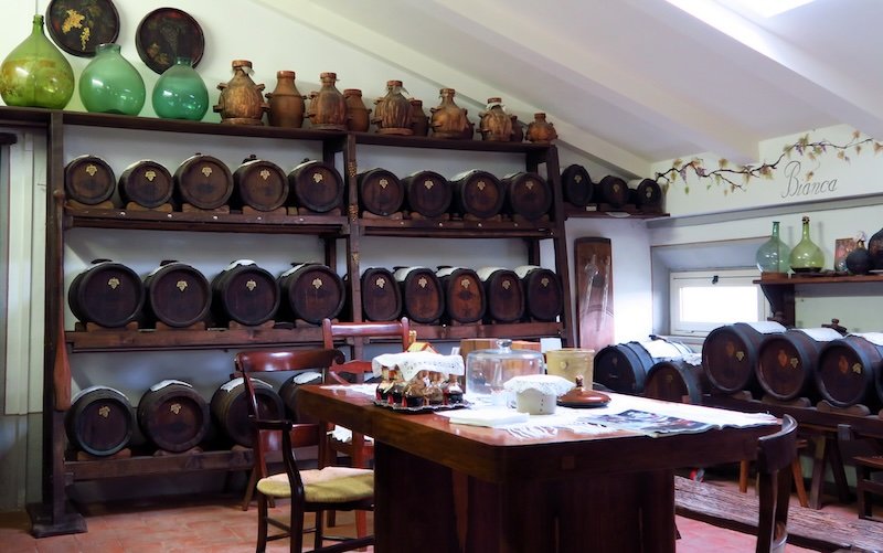 Wooden barrels of traditional balsamic vinegar aging inside an acetaia in Modena, Emilia-Romagna, Italy, with rustic furnishings and glass vessels, capturing the artisanal process and centuries-old craftsmanship behind this iconic Italian delicacy