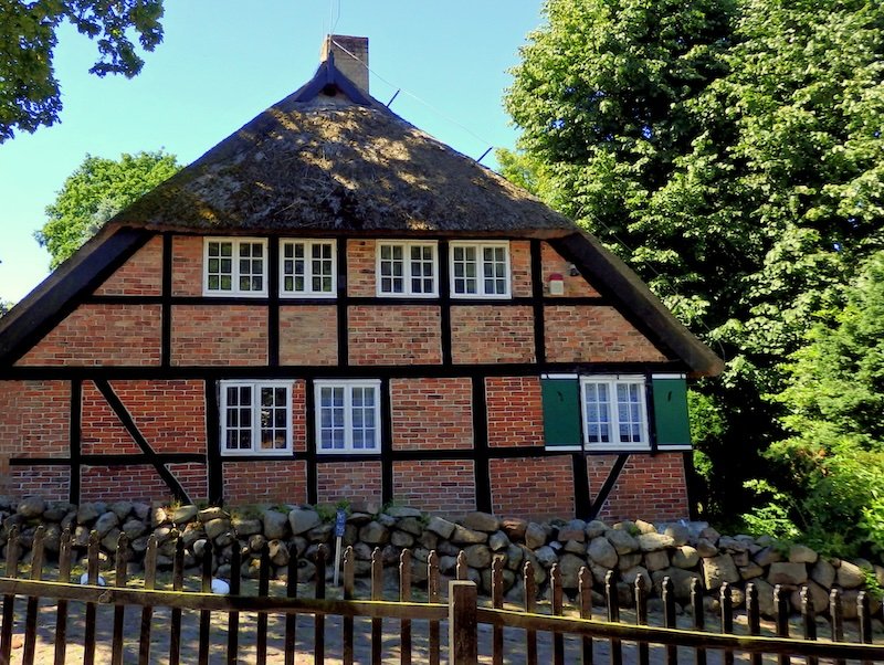 Traditional half-timbered brick house with a thatched roof on Rügen Island, Germany, framed by lush green trees and a rustic stone fence under a clear summer sky