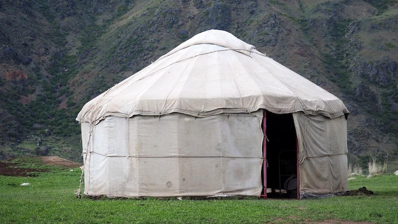 A traditional Kyrgyz yurt with a canvas covering set on a grassy field against a backdrop of rugged green mountains during the 3-Day Panorama Trek in Kyrgyzstan