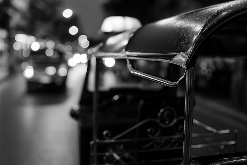 A black and white photograph of a tuk-tuk in Patong Beach, Phuket, taken at night. The chrome handrail and raindrops on the roof are in sharp focus, while the bustling street with lights and vehicles is blurred into bokeh, creating a cinematic scene
