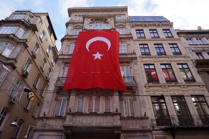 A massive red Turkey national flag with a white crescent and star draped over a historic stone building facade along a bustling street in Istanbul, symbolizing Turkish pride and heritage