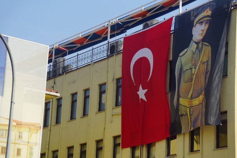 Large Turkey national flag and a military-themed propaganda poster hanging side by side on the facade of a building in Istanbul, symbolizing patriotism and national pride in an urban setting