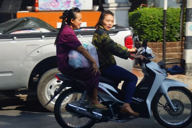 Two ladies riding the same scooter carrying items without helmets in SE Asia 