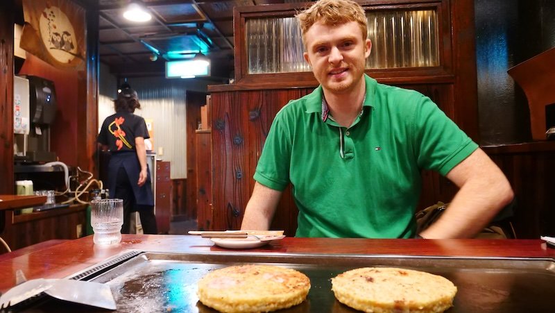 Two okonomiyaki being cooked at the grill on the table in Osaka, Japan with a smiling Nomadic Samuel eagerly waiting in anticipation of a great local meal 