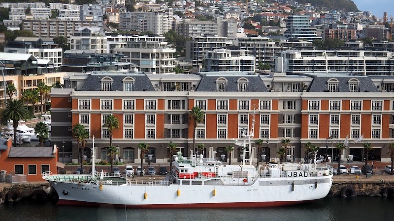 A white ship docked at the bustling V&A Waterfront in Cape Town, South Africa, with elegant colonial-style buildings and modern city apartments rising in the background