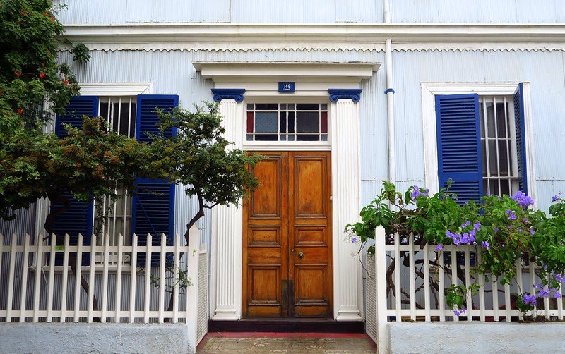 Historic colonial-style house in Valparaíso, Chile, featuring wooden doors, blue shutters, white picket fence, and vibrant flowers, showcasing the city’s unique seaport architecture