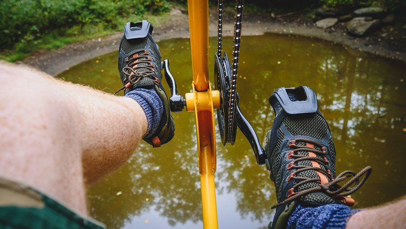Close-up of feet pedalling the bright yellow Vélo Volant canopy bicycle high above a reflective forest pond at Au Diable Vert in Sutton, Quebec, during a treetop adventure in the Eastern Townships