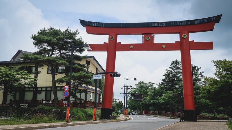 Massive vermilion Torii Gate straddling the road leading toward Lake Chuzenji in Nikko, Japan, with traditional buildings, pine trees, and a cloudy summer sky in the background