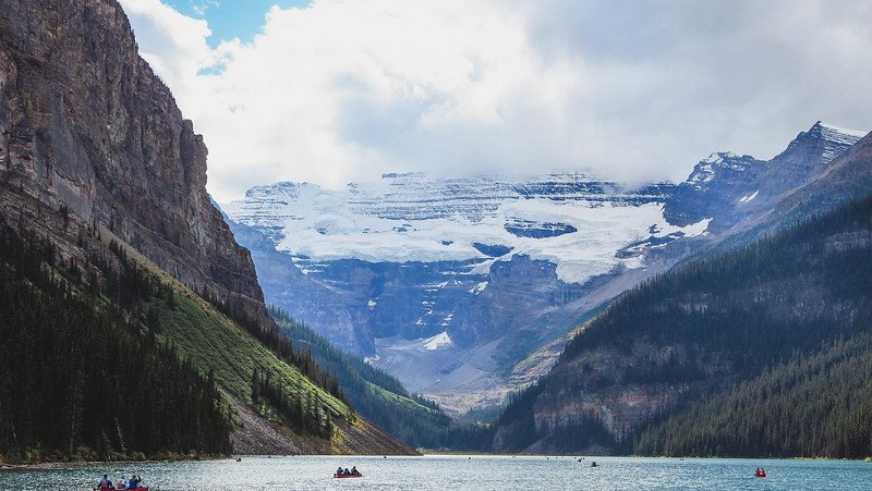 The icy expanse of Victoria Glacier dominating the backdrop above the turquoise waters of Lake Louise, framed by steep forested slopes and rugged cliffs in the Canadian Rockies, with small canoes dotting the lake