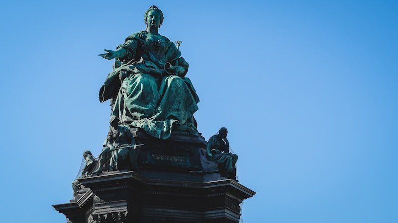 Close-up of the grand Maria Theresa monument in Vienna, Austria, featuring a bronze statue of the empress seated on a throne surrounded by allegorical figures against a clear blue sky Close-up of the grand Maria Theresa monument in Vienna, Austria, featuring a bronze statue of the empress seated on a throne surrounded by allegorical figures against a clear blue sky