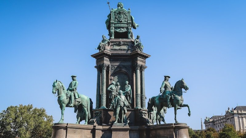 Majestic Maria Theresa monument in Vienna, Austria, showcasing the empress seated atop a grand pedestal surrounded by bronze statues of generals and horsemen under a clear blue sky Majestic Maria Theresa monument in Vienna, Austria, showcasing the empress seated atop a grand pedestal surrounded by bronze statues of generals and horsemen under a clear blue sky