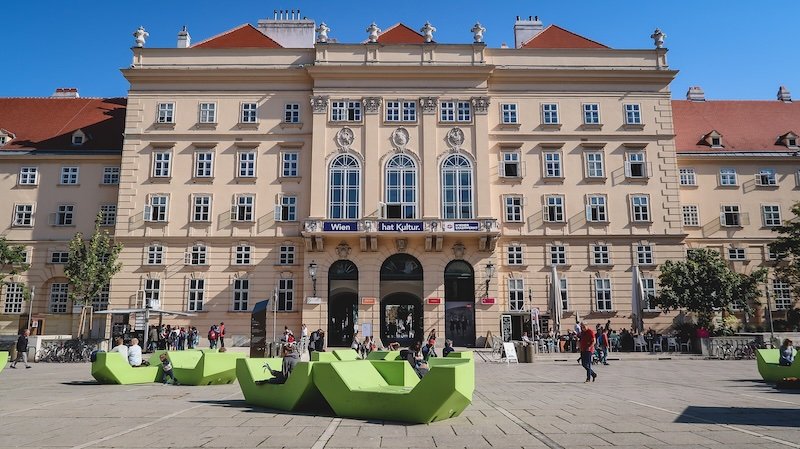 Vibrant courtyard of the MuseumsQuartier in Vienna, Austria, with people relaxing on bright green loungers in front of the grand baroque façade under a sunny blue sky Vibrant courtyard of the MuseumsQuartier in Vienna, Austria, with people relaxing on bright green loungers in front of the grand baroque façade under a sunny blue sky