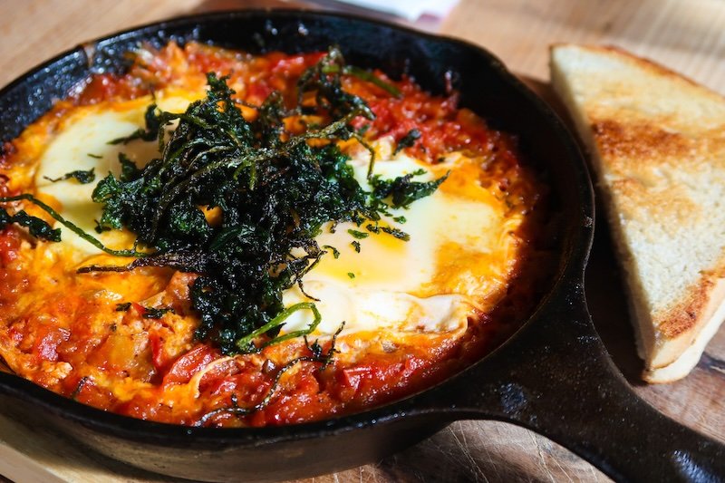 Close-up of a sizzling skillet of Shakshuka served in Vienna, Austria, featuring poached eggs in spiced tomato sauce topped with crispy herbs and a slice of toasted bread on the side Close-up of a sizzling skillet of Shakshuka served in Vienna, Austria, featuring poached eggs in spiced tomato sauce topped with crispy herbs and a slice of toasted bread on the side