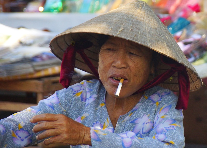 Vietnamese vendor smoking a cigarette and wearing a conical hat in Hoi Ai, Vietnam Vietnamese vendor smoking a cigarette and wearing a conical hat in Hoi Ai, Vietnam