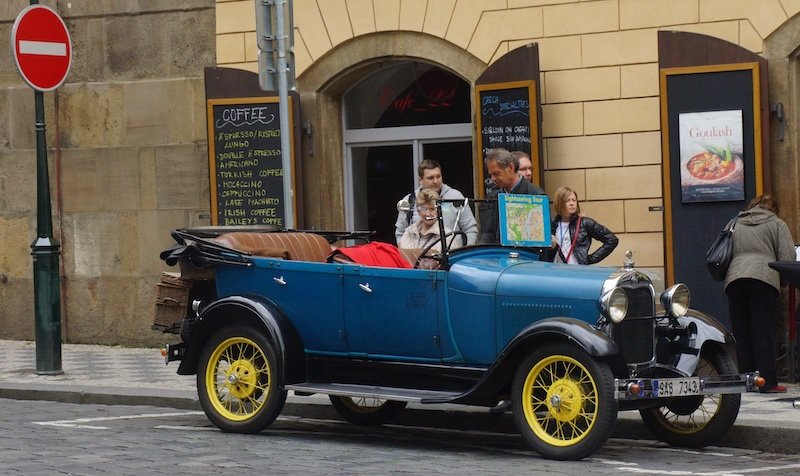 Vintage blue convertible car with yellow wheels parked on a cobblestone street offering guided sightseeing tours in Prague, Czech Republic, with tourists gathered around