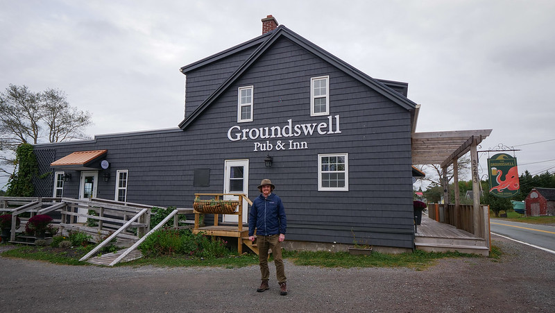 Visiting The Groundswell Pub & Inn in Isle Madame, Cape Breton with Nomadic Samuel standing outside of the building in Canada