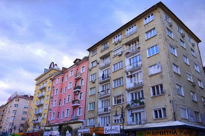 Vitosha Boulevard in Sofia, Bulgaria, featuring pastel-colored and Soviet-style apartment buildings with shops, cafés, and restaurants at street level under a cloudy sky, capturing the mix of architecture and lively atmosphere of the city’s main street