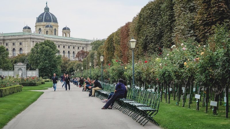 People relaxing on benches along a rose-lined path in Volksgarten with the grand dome of the Kunsthistorisches Museum visible in the background in Vienna, Austria People relaxing on benches along a rose-lined path in Volksgarten with the grand dome of the Kunsthistorisches Museum visible in the background in Vienna, Austria