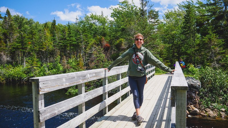 That Backpacker Audrey Bergner smiles while standing on a wooden bridge surrounded by lush green forest and calm waters during a summer visit to Warren Lake in Cape Breton Highlands National Park, a scenic stop along the Cabot Trail in Nova Scotia