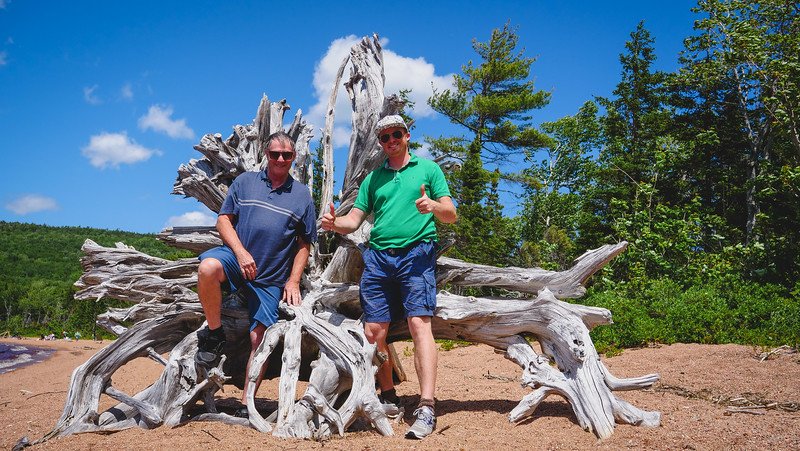Nomadic Samuel and his father George pose smiling beside a massive weathered driftwood tree root on the sandy shore of Warren Lake in Cape Breton Highlands National Park, surrounded by lush green forest and blue skies during their scenic road trip along the Cabot Trail in Nova Scotia