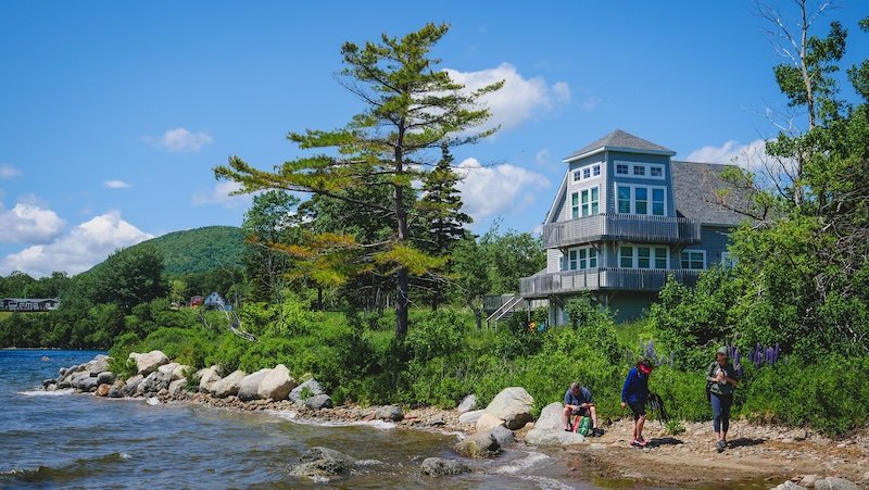 Travellers explore the rocky shoreline beside a modern waterfront house surrounded by lush greenery and forested hills under blue skies along the scenic Cabot Trail in Cape Breton, Nova Scotia