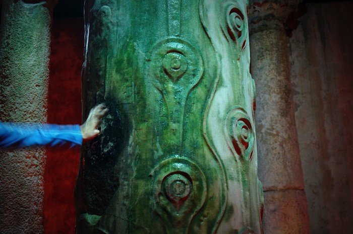 A close-up of the legendary Weeping Column inside the Basilica Cistern in Istanbul, Turkey, showing intricate teardrop carvings glistening with moisture as a visitor touches the worn spot believed to bring good luck