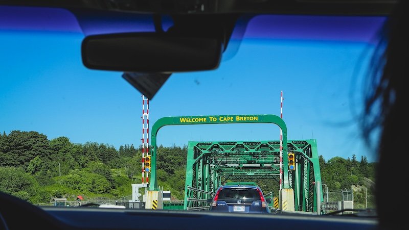 A view from inside a car approaching the green steel bridge with the bright yellow “Welcome to Cape Breton” sign, marking the gateway to Nova Scotia’s Cabot Trail on a sunny summer road trip