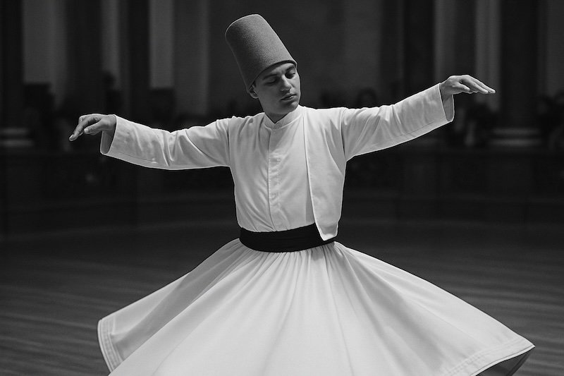 A black and white photograph of a Whirling Dervish mid-spin during a Sema ceremony in Istanbul, Turkey. His gaze is softly fixed on his left thumb, demonstrating the spinning technique. The flowing robe flares around him against a softly blurred hall background A black and white photograph of a Whirling Dervish mid-spin during a Sema ceremony in Istanbul, Turkey. His gaze is softly fixed on his left thumb, demonstrating the spinning technique. The flowing robe flares around him against a softly blurred hall background