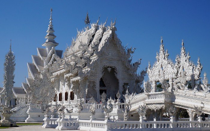 Wat Rong Khun, famously known as the White Temple in Chiang Rai, Thailand, gleaming under a clear blue sky. This intricate contemporary Buddhist temple features ornate carvings, mirror mosaics, and elaborate white structures that symbolize purity