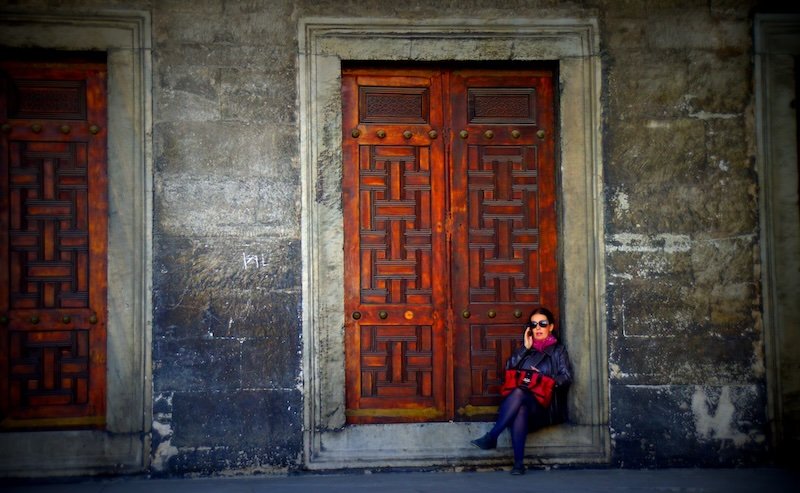 Woman in dark clothing relaxing against an intricately carved wooden door framed by stone at Blue Mosque in Istanbul, Turkey, showcasing traditional Ottoman architectural details