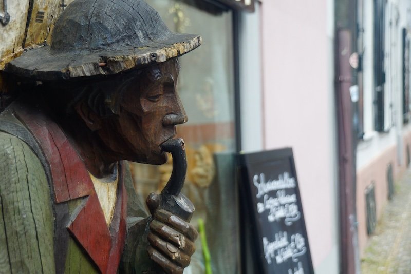 Close-up of a weathered wooden carving of a man in a hat smoking a pipe, displayed outside a traditional shop in Freiburg im Breisgau’s old town
