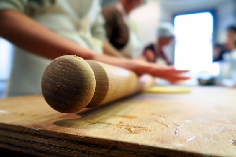 Close-up of a wooden rolling pin on a flour-dusted board during a pasta-making class in Emilia-Romagna, Italy, with blurred figures shaping dough in the background, highlighting the hands-on culinary traditions of authentic Italian cooking