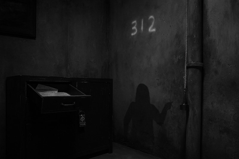 A black and white photograph of a dimly lit zombie-themed escape room featuring classic puzzle archetypes: an open drawer filled with maps, a cabinet with a directional lock, faint UV numbers glowing “312” on the wall, and a string with a magnet hanging near a pipe. The moody lighting adds mystery